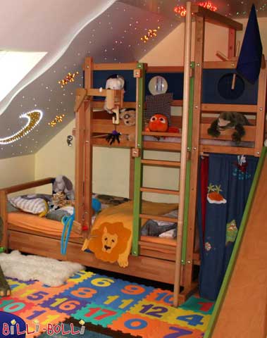 Etagenbett-seitlich-versetzt in Buche, Leiterposition B über der unteren Schlafebene. Außerdem zu sehen: Bettkastenbett, Feuerwehrstange, Spielkran, Rutsche und farbig lackierte Bullaugen-Themenbretter.